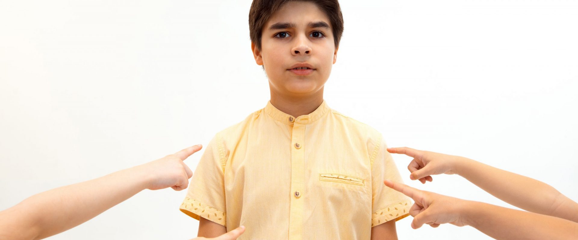 Little boy standing alone and suffering an act of bullying while children mocking in the background. Sad young schoolboy standing on studio against white background.